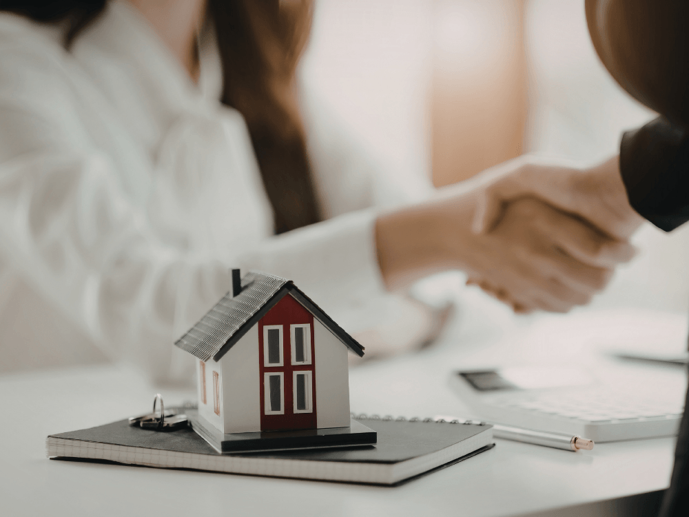 Miniatura de casa sobre caderno enquanto duas pessoas apertam as mãos ao fundo em uma negociação imobiliária.