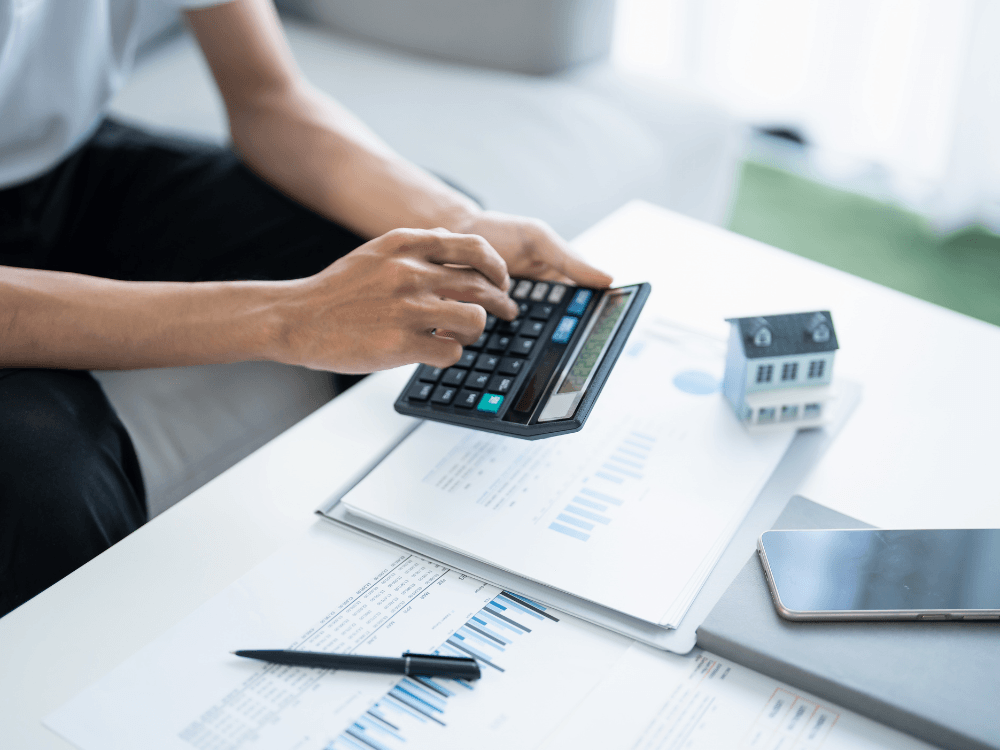 Pessoa utiliza calculadora ao lado de documentos e maquete de casa para mostrar como precificar imóvel