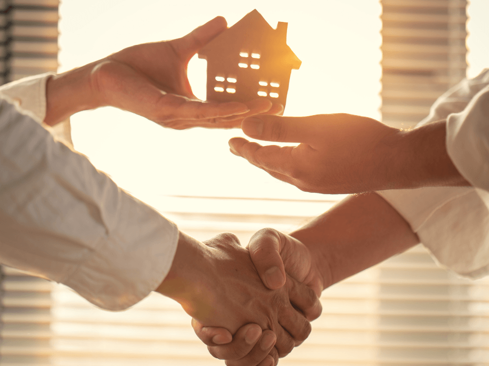 Duas pessoas apertam as mãos enquanto uma miniatura de casa é entregue, simbolizando a negociação após definir o valor do imóvel.